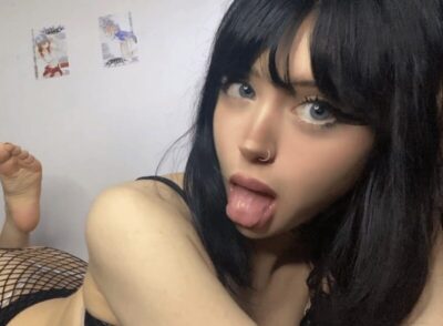 Woman with black hair and blue eyes sticking out her tongue, nose piercing, wearing a black top, lying on a surface with a fishnet garment visible.