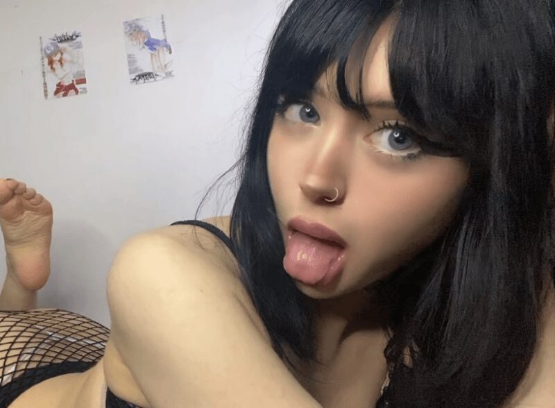 Woman with black hair and blue eyes sticking out her tongue, nose piercing, wearing a black top, lying on a surface with a fishnet garment visible.