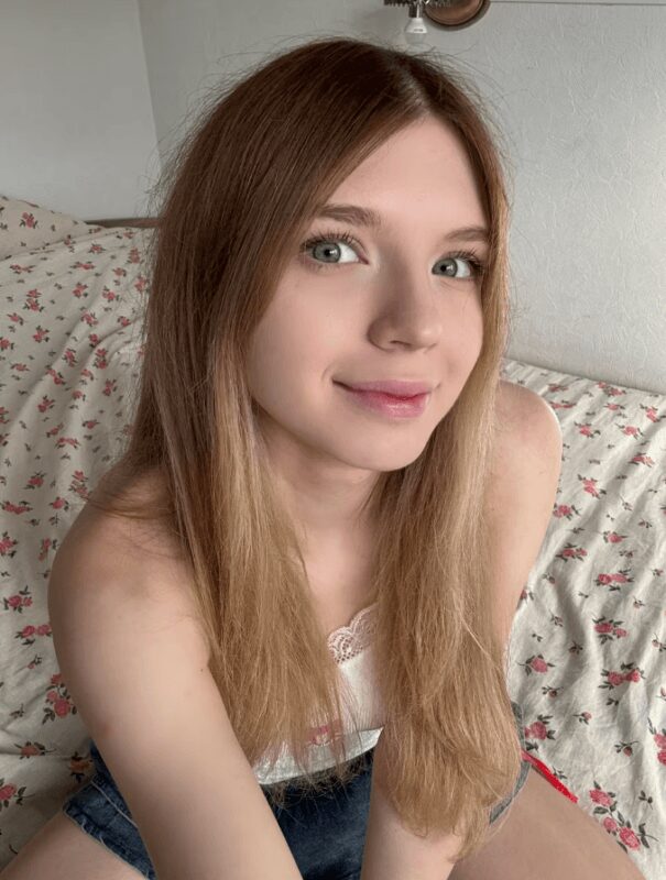 Young woman with long light brown hair and blue eyes smiling at the camera, seated on a floral bedspread.