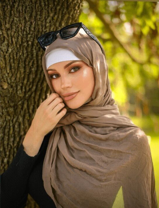 Woman wearing a beige hijab and white undercap, with sunglasses resting on her head, outdoors by a tree with greenery in the background.