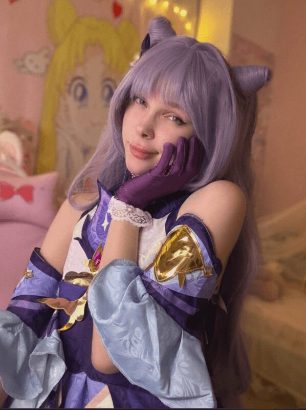 Cosplay of a woman with lavender wig and horn headband, wearing a detailed purple and light blue costume with gloves, posing with hand on her face in a pink-themed room.
