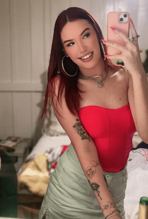 Young woman taking a mirror selfie in a bedroom, wearing a red strapless top, light green skirt, hoop earrings, and layered necklaces; visible tattoos on her arm with a pink phone in her hand.