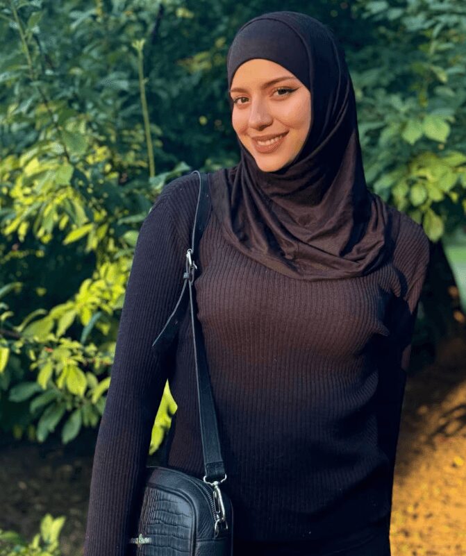 Smiling woman in a black hijab and sweater standing outdoors with greenery, carrying a black crossbody bag.