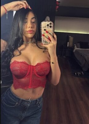 Self-portrait of a woman in a red lace bustier taking a mirror selfie with her phone.