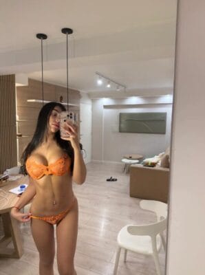 Woman in orange lace lingerie taking a mirror selfie in a modern living room with a beige sofa.