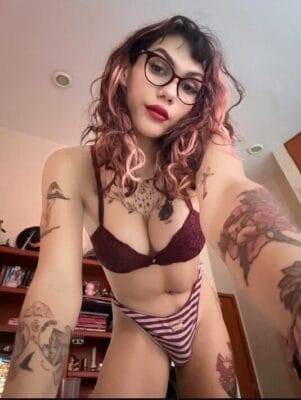 Person in burgundy lace bra and striped panties posing for a selfie, with pink-tinted hair and glasses, tattoos visible on arms and chest in a bedroom.
