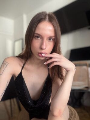 Portrait of a woman with long brown hair wearing a black sequined halter top, indoors, with one hand near her lips.