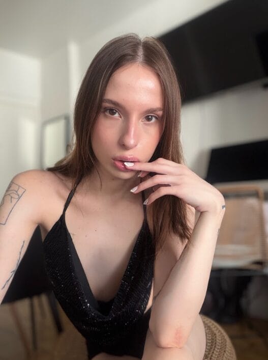 Portrait of a woman with long brown hair wearing a black sequined halter top, indoors, with one hand near her lips.