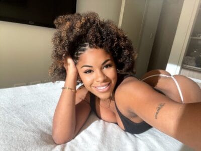 Smiling woman with curly hair lying on a bed, propped on her arm, taking a selfie in a sunlit room wearing a black top and white bottoms (selfie).