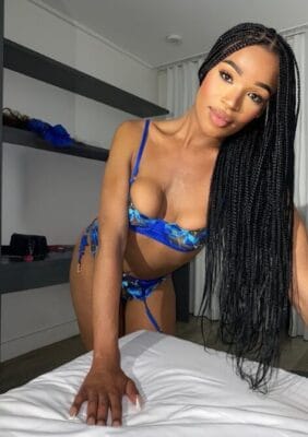 Woman in a blue floral lingerie set leaning forward on a bed in a bedroom, posing for a selfie-style photo.
