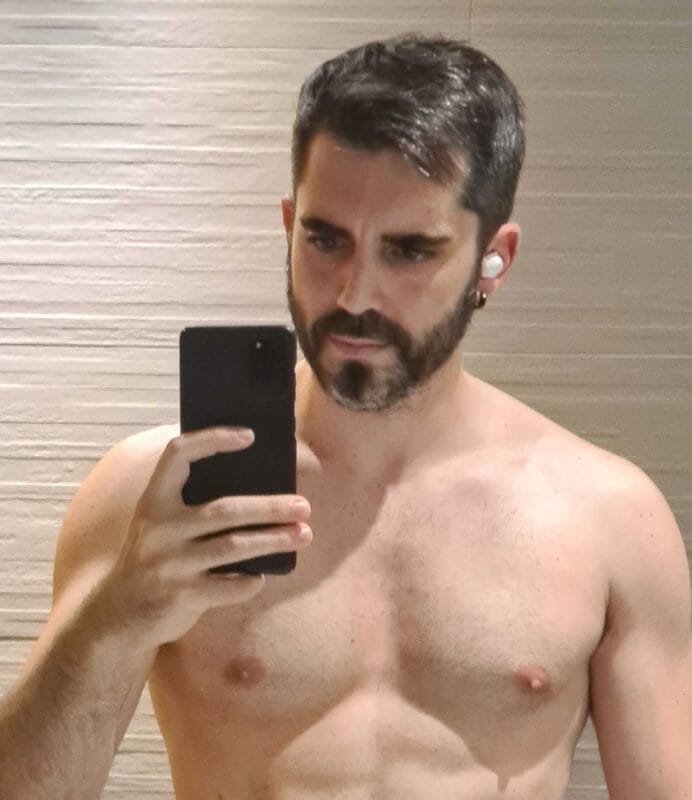 Shirtless man with dark hair and beard taking a mirror selfie in a bathroom, holding a black smartphone in his left hand.