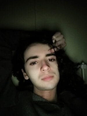 Young man with dark hair relaxing in bed, hand in his hair, dim bedroom lighting on his face.