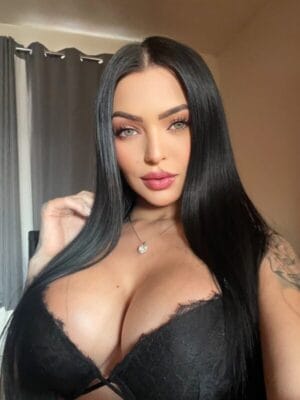 Close-up selfie of a woman with long black hair, wearing a black lace bra and a pendant necklace, indoors.