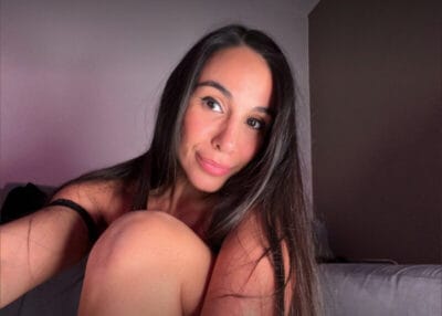 Close-up selfie of a smiling woman with long dark hair, leaning toward the camera in a softly lit room on a couch.