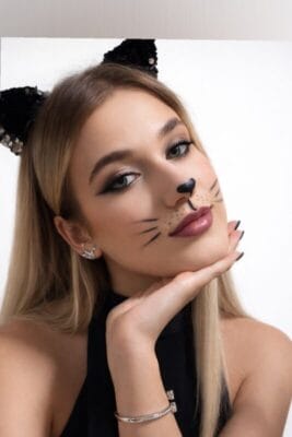 Woman with cat makeup and ears resting her chin on her hand, wearing a black top and silver jewelry, posed against a white background.