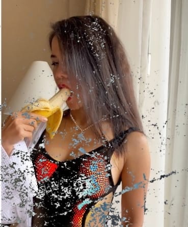 Woman in a sequined black dress with red detailing by a window, holding a peeled banana near her mouth.